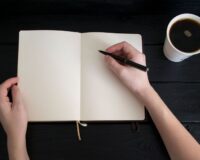 Hand schrijft met een zwarte pen in een leeg notitieboek naast koffie