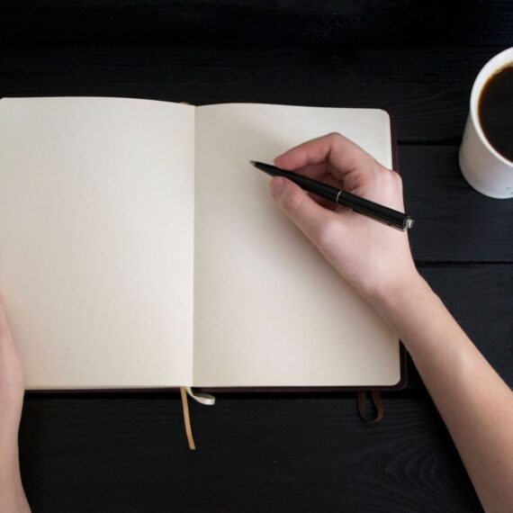 Hand schrijft met een zwarte pen in een leeg notitieboek naast koffie