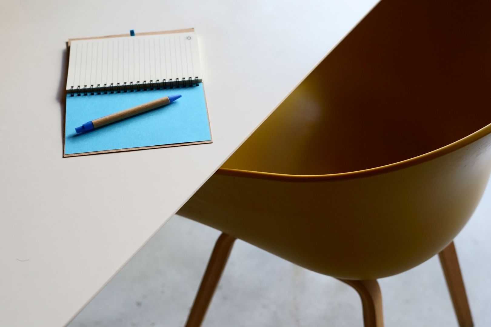 Blauw notitieboek met pen op een witte tafel naast een gele stoel